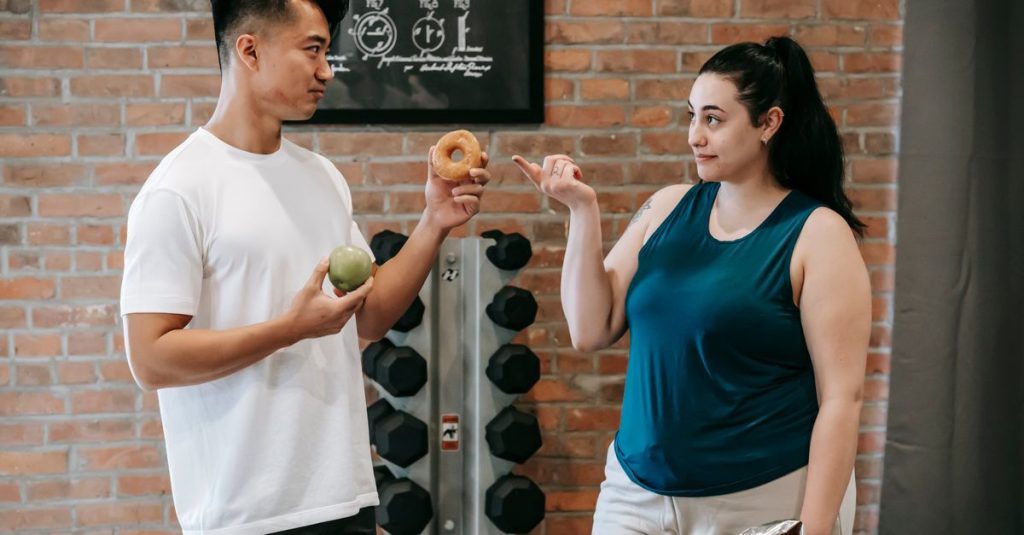 Side view of Asian personal instructor with apple and doughnut offering healthy food to plus size woman while standing in gym