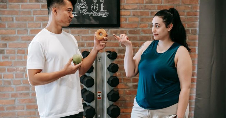Side view of Asian personal instructor with apple and doughnut offering healthy food to plus size woman while standing in gym