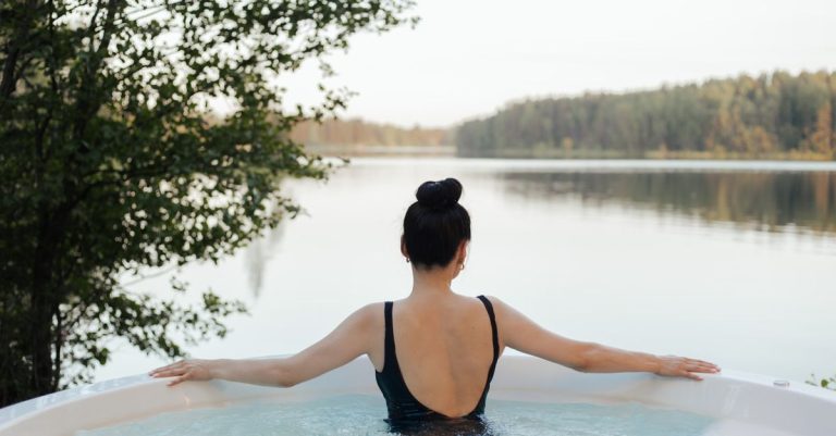 A woman relaxes in a jacuzzi outdoors, overlooking a serene lake and forest landscape.