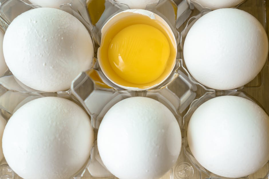 A high-angle view of fresh eggs in a carton with one cracked open showing the yolk.