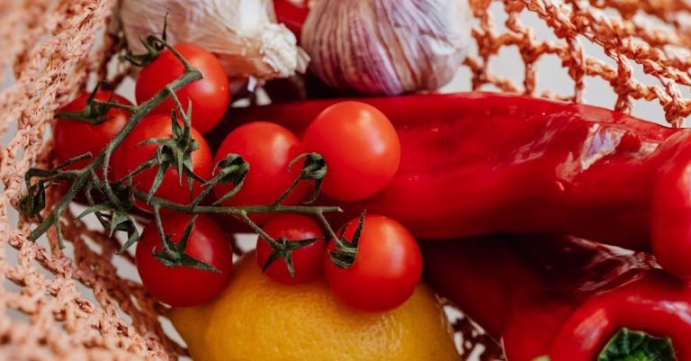 Top view of ripe tomatoes on branch arranged amidst fresh yellow lemon and red chili peppers with garlic in pink food net