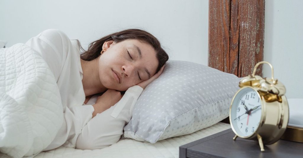 Young calm female with dark hair lying on side with hand under head on white comfortable bed under blanket while sleeping in morning time