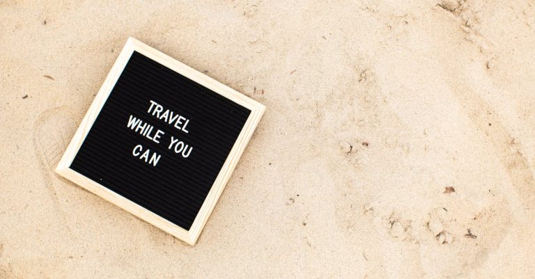 Letter board with 'Travel While You Can' message displayed on sandy beach scene, inspiring wanderlust.