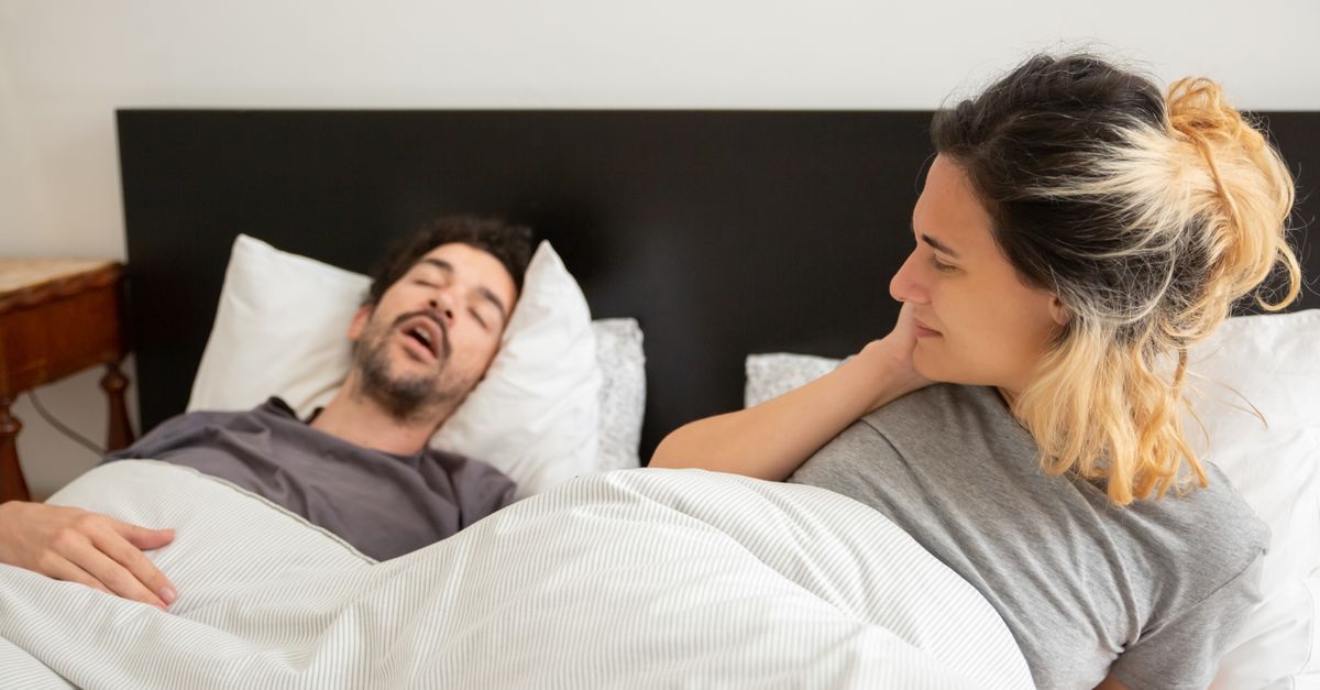 A couple in bed with a man snoring loudly and a woman looking restless on her pillow.