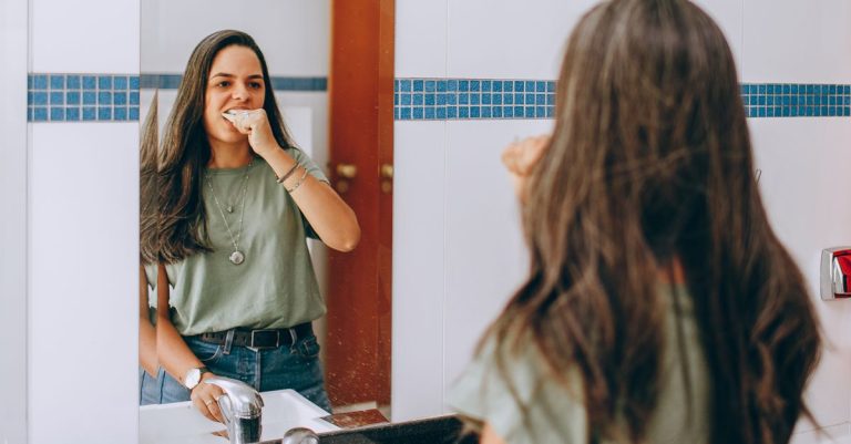 A young woman brushes her teeth, looking in the bathroom mirror. Self-care and hygiene concept.