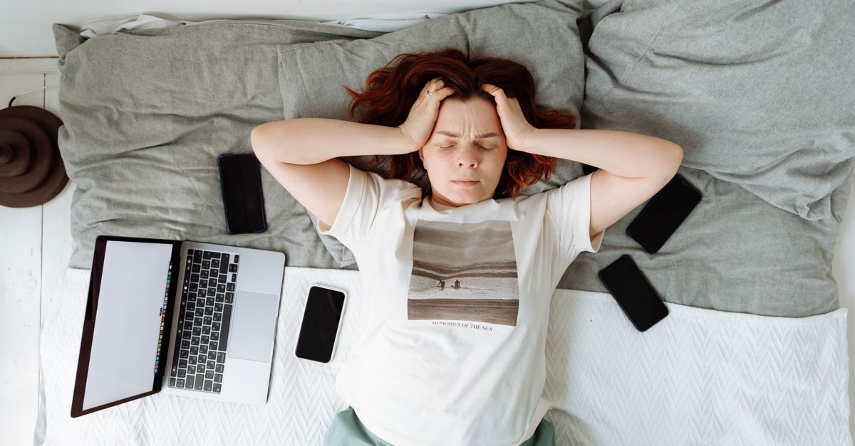 A distressed woman lying in bed surrounded by digital devices, showing signs of stress.