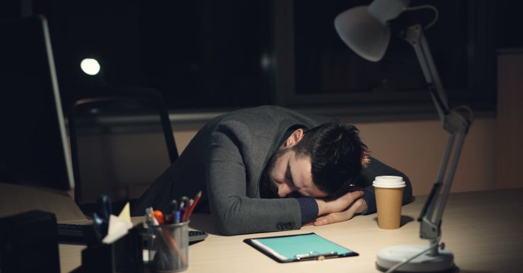 Man in office suit sleeping on desk with coffee and tablet nearby, symbolizing work fatigue.