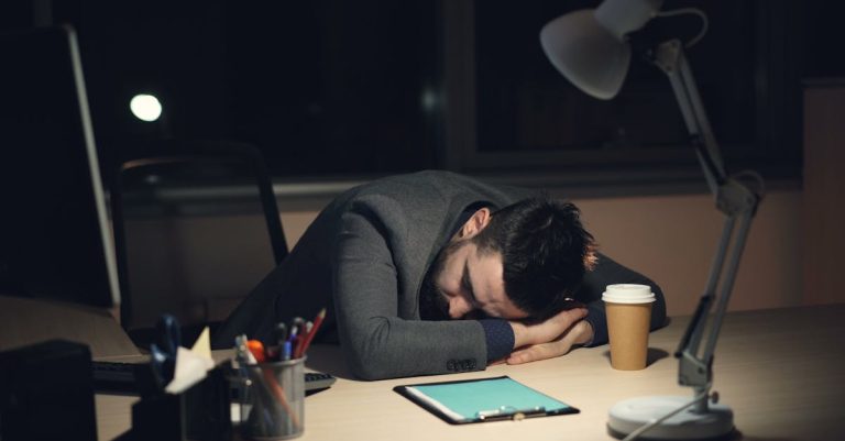 Man in office suit sleeping on desk with coffee and tablet nearby, symbolizing work fatigue.
