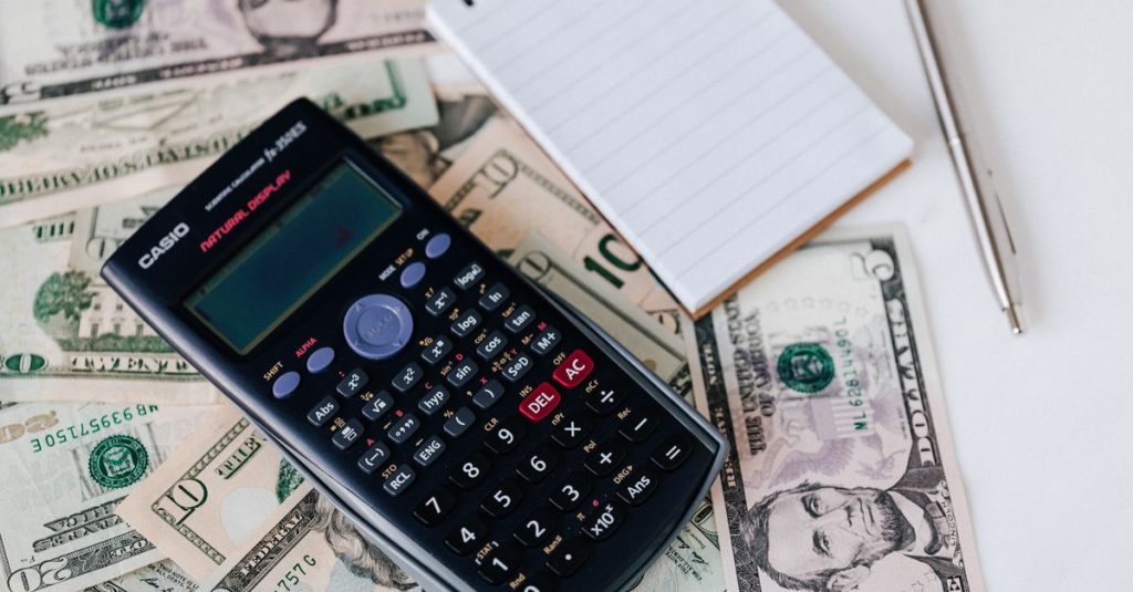 From above electronic calculator and notepad placed over United States dollar bills together with metallic pen for budget planning and calculation