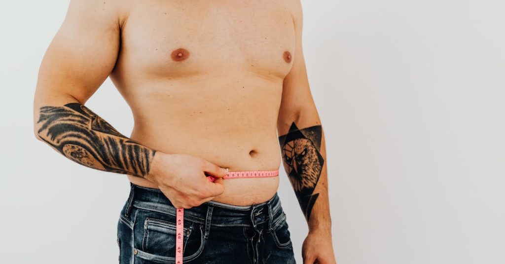 Shirtless man using measuring tape to check waist size, showcasing tattoos for fitness concept.