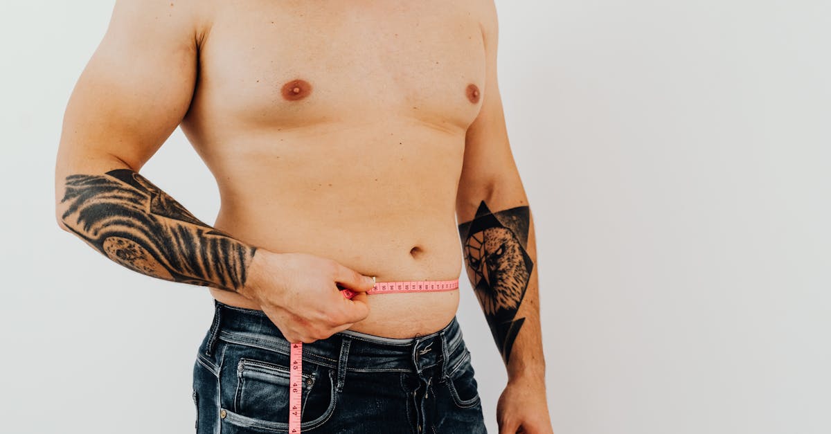 Shirtless man using measuring tape to check waist size, showcasing tattoos for fitness concept.