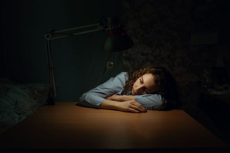 A woman in a dimly lit room rests her head on a desk, illuminated by a desk lamp.